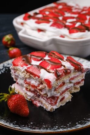 A slice of strawberry icebox cake topped with sliced strawberries and chocolate drizzle on a black plate.