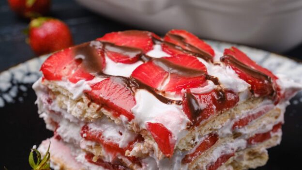A slice of strawberry icebox cake topped with sliced strawberries and chocolate drizzle on a black plate.