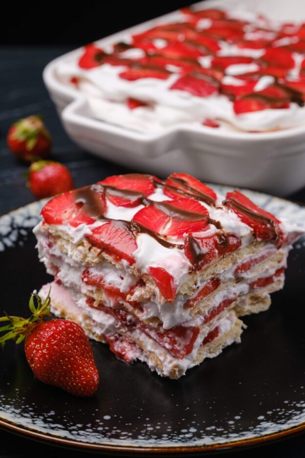 A slice of strawberry icebox cake topped with sliced strawberries and chocolate drizzle on a black plate.