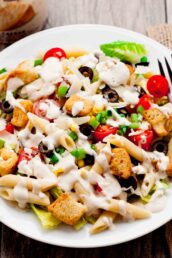 A plate of Caesar pasta salad with penne, cherry tomatoes, black olives, croutons, green onions, and lettuce, topped with a creamy dressing.