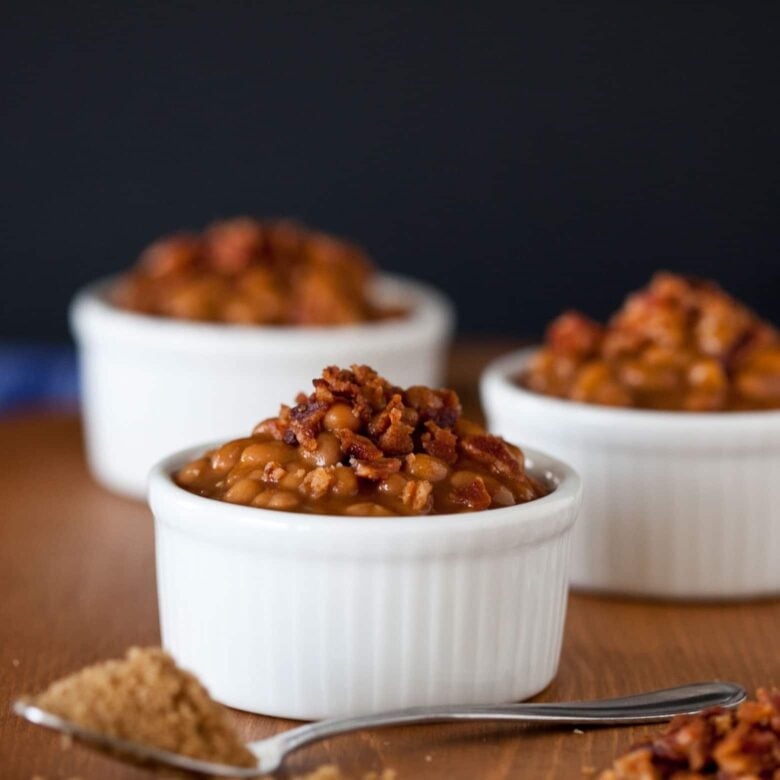 Three white ramekins filled with brown sugar baked beans topped with bacon bits on a wooden surface.