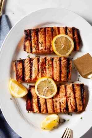 Three grilled salmon fillets with perfect grill marks are served on a white plate, garnished with lemon slices and chopped herbs - an Easy Grilled Salmon dinner that’s both delicious and simple to prepare.