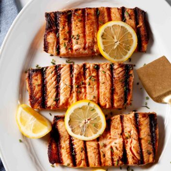 Three grilled salmon fillets with perfect grill marks are served on a white plate, garnished with lemon slices and chopped herbs - an Easy Grilled Salmon dinner that’s both delicious and simple to prepare.