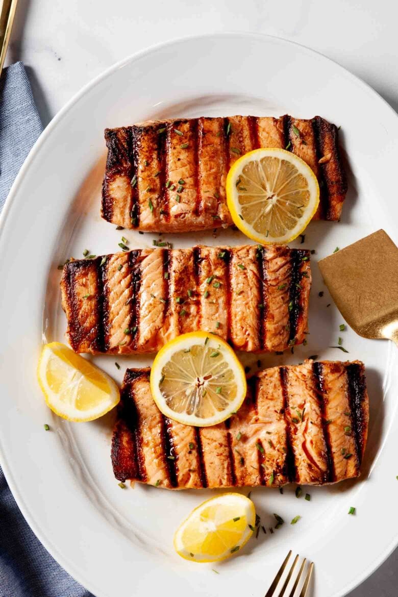 Three grilled salmon fillets with perfect grill marks are served on a white plate, garnished with lemon slices and chopped herbs - an Easy Grilled Salmon dinner that’s both delicious and simple to prepare.
