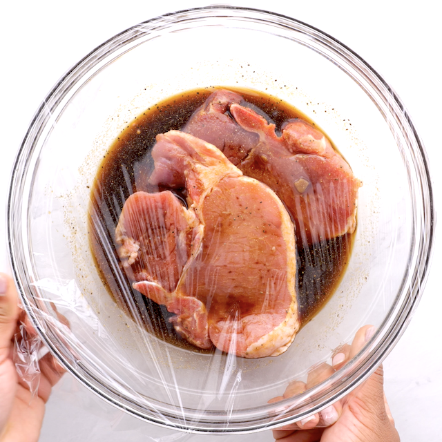 Pork chops with marinade in a glass bowl covered with plastic wrap.