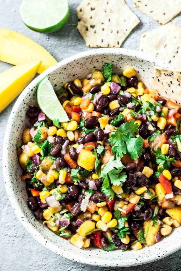 Colorful mango salad in a speckled bowl featuring black beans, corn, diced vegetables, herbs, and a lime wedge.