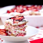 A close-up of a slice of strawberry icebox cake on a white plate, topped with sliced strawberries and drizzled with chocolate.
