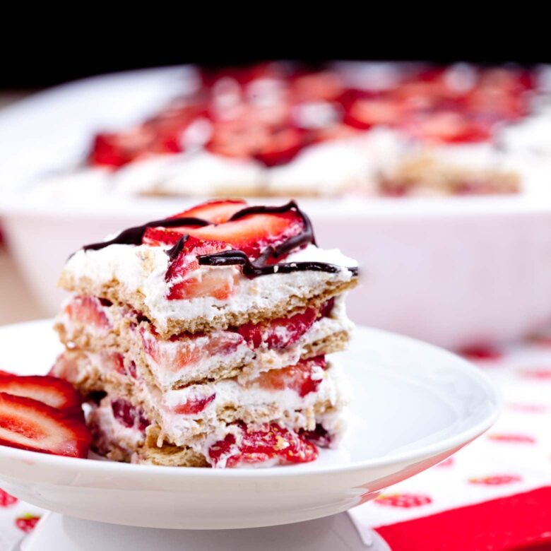 A close-up of a slice of strawberry icebox cake on a white plate, topped with sliced strawberries and drizzled with chocolate ganache. 