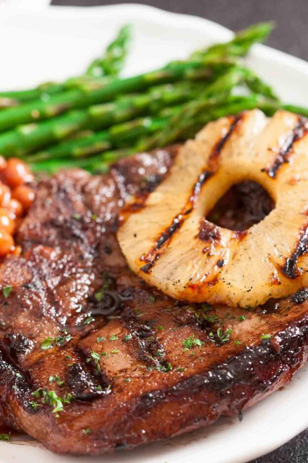 A plate with grilled pork chops with marinade topped with a grilled pineapple slice, accompanied by baked beans and asparagus.
