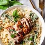 A plate of healthy chicken Alfredo in creamy sauce topped with sliced grilled chicken, fresh basil leaves, and grated cheese.