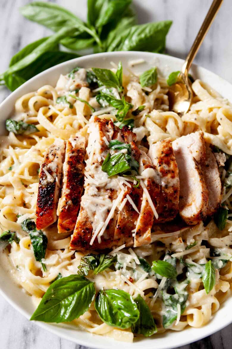 Closeup shot of a plate of healthy chicken Alfredo with sliced grilled chicken, fresh basil leaves, and grated cheese.