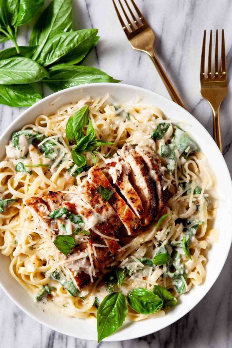 A plate of healthy chicken Alfredo in creamy sauce topped with sliced grilled chicken, fresh basil leaves, and grated cheese.