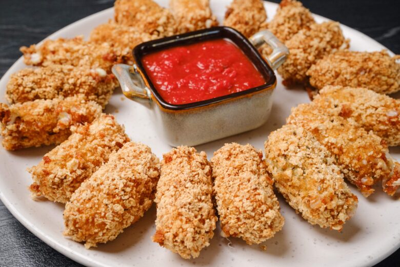 A white plate with an arranged ring of breaded mozzarella sticks around a square dish of marinara sauce.