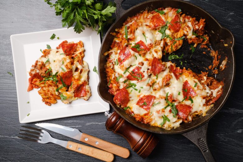 A skillet filled with pizza pasta topped with melted cheese and pepperoni, garnished with fresh herbs. A portion is served on a white plate.