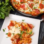 A portion of pizza pasta with melted cheese, pepperoni, ground meat, and herbs on a white plate, with a skillet containing the same dish in the background.