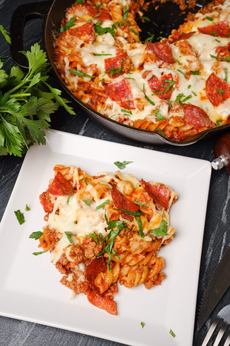 A portion of pizza pasta with melted cheese, pepperoni, ground meat, and herbs on a white plate, with a skillet containing the same dish in the background.
