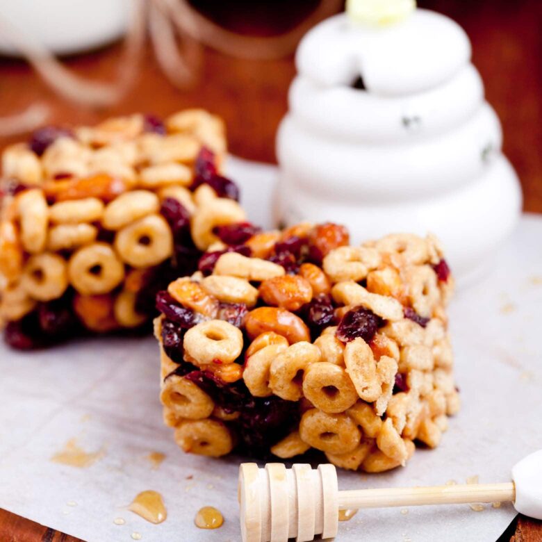 Two square-shaped cereal bars made with cheerios and dried fruits, placed on a piece of parchment paper.