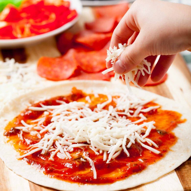 A person sprinkling shredded cheese over pizza sauce on a tortilla, with pepperoni and sliced vegetables in the background.