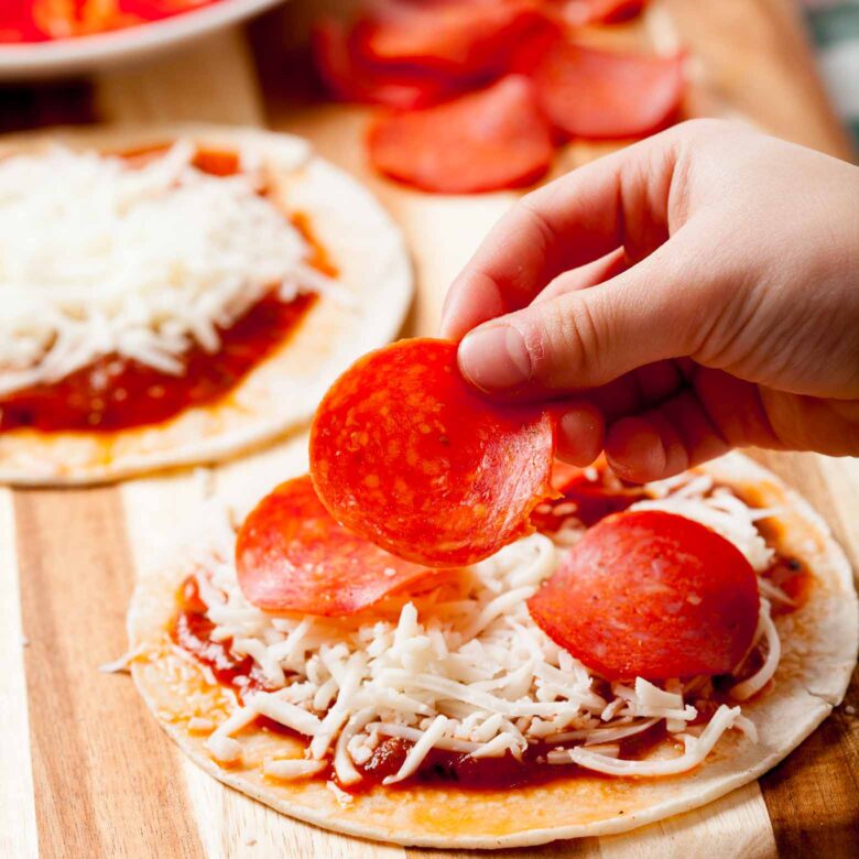 A person placing slices of pepperoni on tortillas with layers of cheese and pizza sauce.