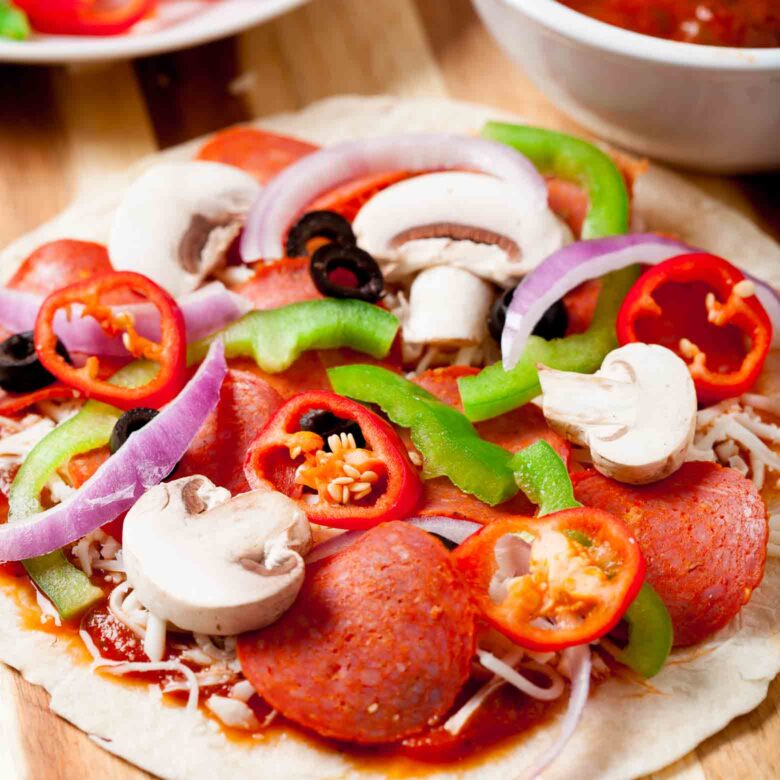 A close-up of a pizzadilla topped with pepperoni, mushrooms, red bell pepper slices, green bell pepper slices, red onion slices, black olives, and cheese on a wooden surface.