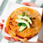 A plate of stacked pizza quesadillas filled with melted cheese, pepperoni, mushrooms, and bell peppers, topped with sliced avocado and cilantro.