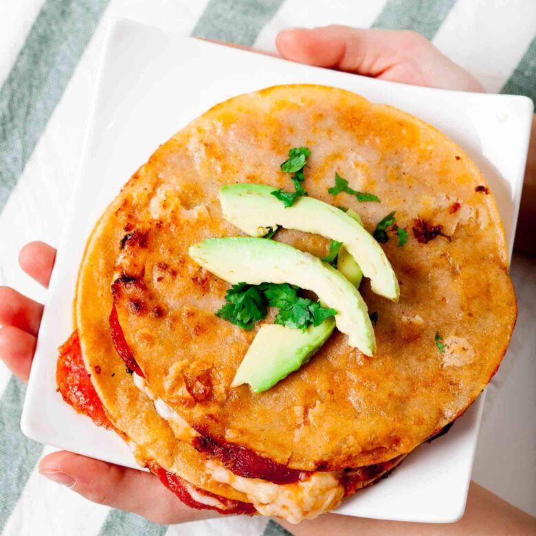 A plate of stacked pizza quesadillas filled with melted cheese, pepperoni, mushrooms, and bell peppers, topped with sliced avocado and cilantro.