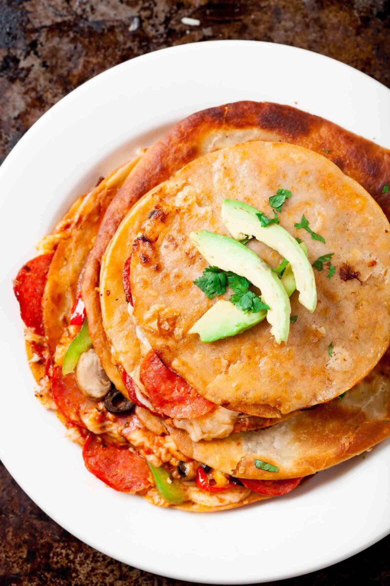 A plate of stacked pizza quesadillas filled with melted cheese, pepperoni, mushrooms, and bell peppers, topped with sliced avocado and cilantro.