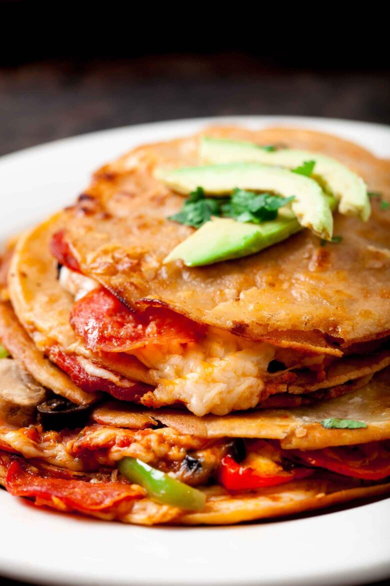 Closeup shot of a plate of stacked quesadillas filled with melted cheese, sautéed vegetables, and topped with avocado slices and cilantro.