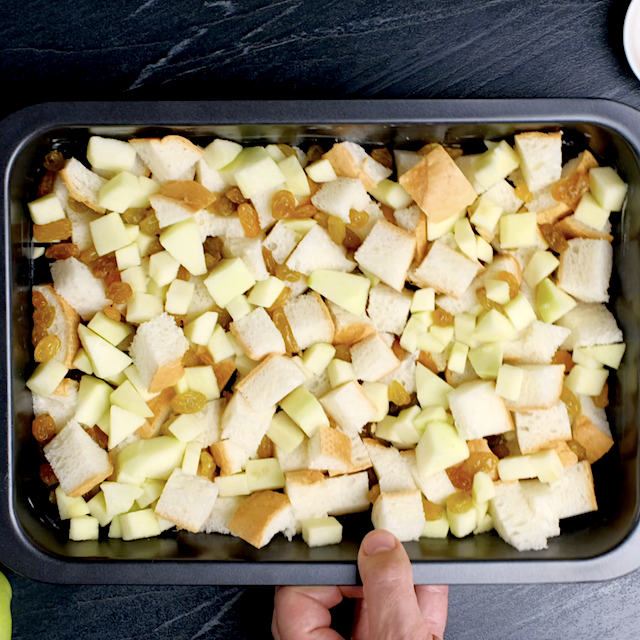 A hand holding a baking dish filled with a mixture of cubed bread, diced apples, and raisins.