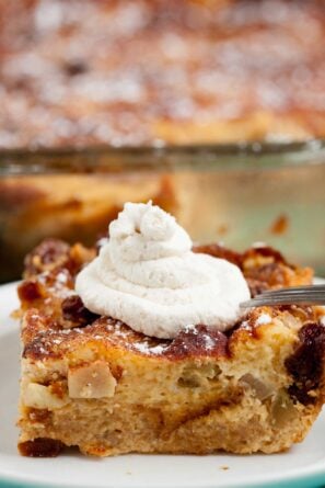 A close-up of a slice of apple bread pudding on a white plate, topped with whipped cream and powdered sugar.