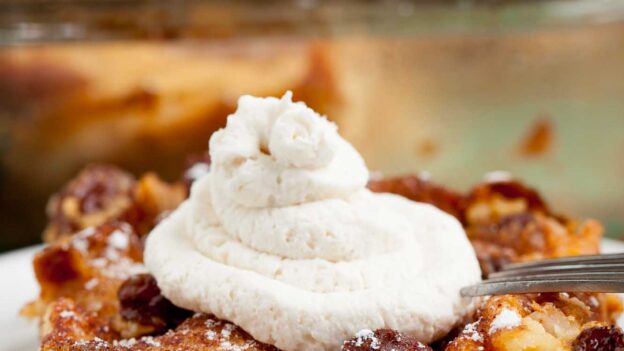A close-up of a slice of apple bread pudding on a white plate, topped with whipped cream and powdered sugar.