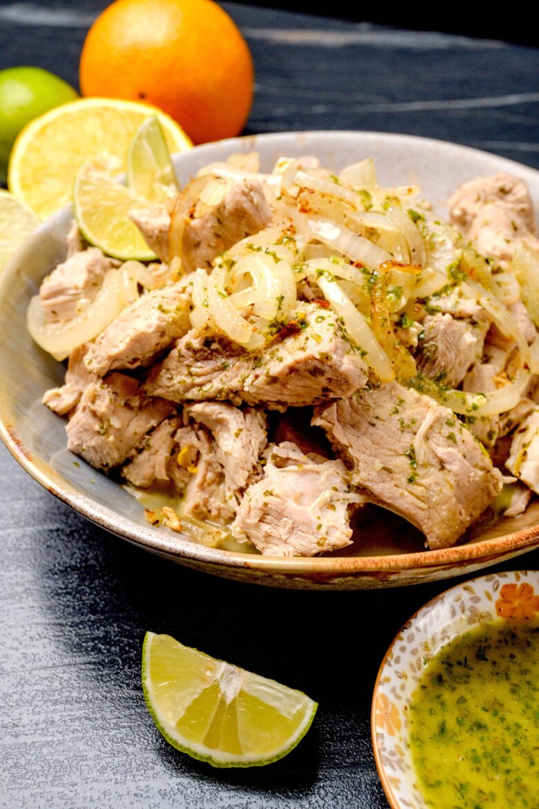 A bowl of Cuban mojo pork topped with onions and surrounded by lime wedges and an orange. 