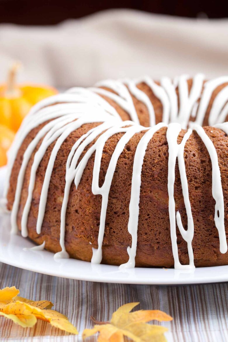 Pumpkin bundt cake with cream cheese glaze on top on a white plate with fall leaves around it.
