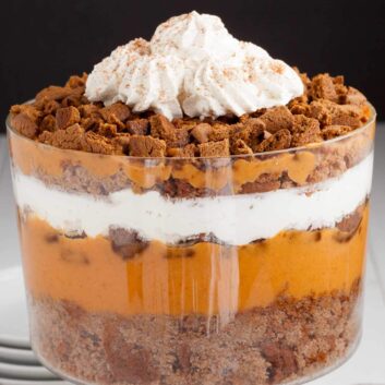 A layered pumpkin trifle dessert in a glass trifle dish with pumpkin pudding mix, whipped cream, and crumbled gingersnap cookies.