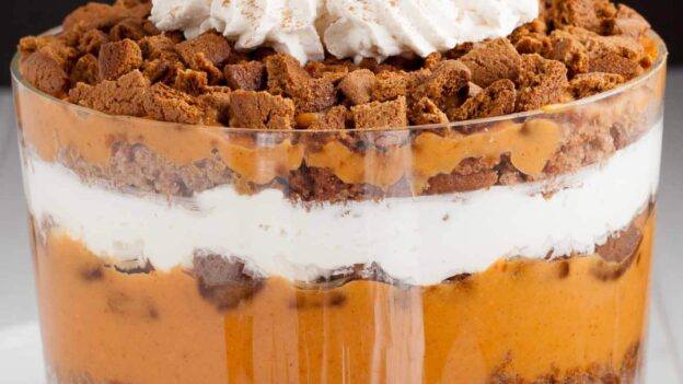 A layered pumpkin trifle dessert in a glass trifle dish with pumpkin pudding mix, whipped cream, and crumbled gingersnap cookies.