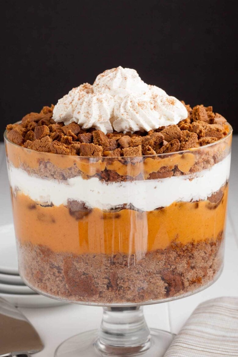 A layered pumpkin trifle dessert in a glass trifle dish with pumpkin pudding mix, whipped cream, and crumbled gingersnap cookies.