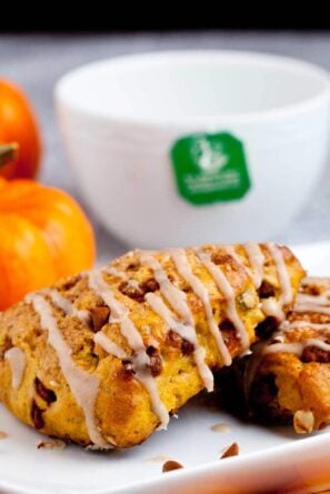 Pumpkin scones drizzled with icing on a white plate, with small pumpkins and a white mug in the background.