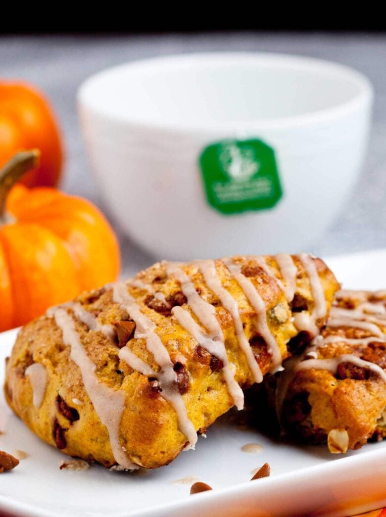 Pumpkin scones drizzled with icing on a white plate, with small pumpkins and a white mug in the background.