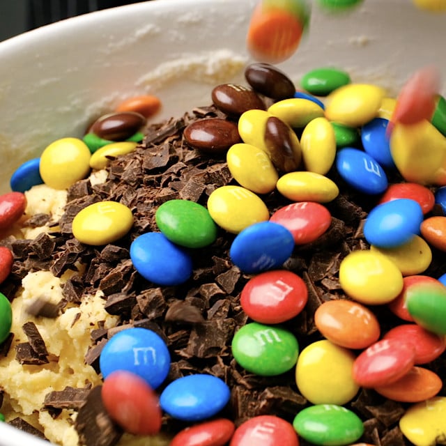 M&M candies being added to cookie dough mixture that also contains chocolate chips to make M&M chocolate chip cookies.