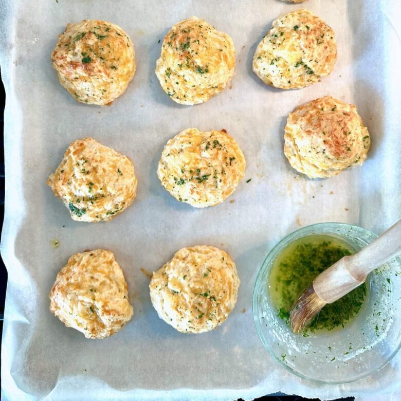 Freshly baked cheddar bay biscuits with herb topping on parchment paper, next to a bowl of herb butter and a brush.