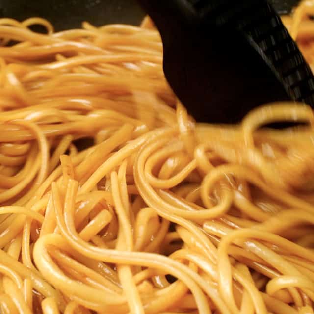 Cooked noodles being tossed with sauces and seasonings to make chicken lo mein.