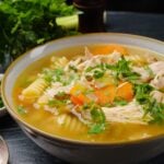 A bowl of chicken noodle soup with carrots, rotini pasta, and chopped herbs.