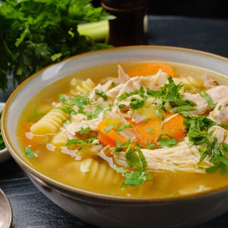 A bowl of chicken noodle soup with carrots, rotini pasta, and chopped herbs. 