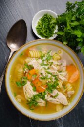 Overhead shot of a bowl of chicken noodle soup with carrots and herbs on a dark surface.