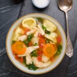 Overhead shot of a bowl of chicken tortellini soup with carrots, kale, and diced tomatoes, with a soup spoon on the side.