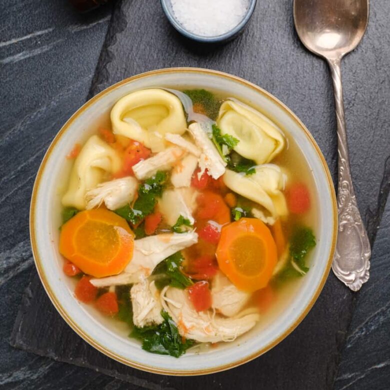 Overhead shot of a bowl of chicken tortellini soup with carrots, kale, and diced tomatoes, with a soup spoon on the side.