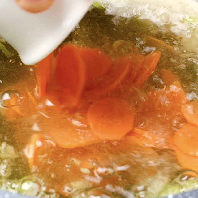 Carrots being added to pot with broth and other veggies to make chicken tortellini soup.