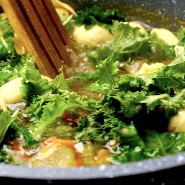 Fresh kale being added alongside frozen tortellini to a pot with broth and veggie mixture to make chicken tortellini soup.