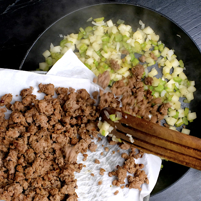 Cooked sausage being added to skillet with sautéed onons, celery, and apples to make cornbread sausage stuffing.
