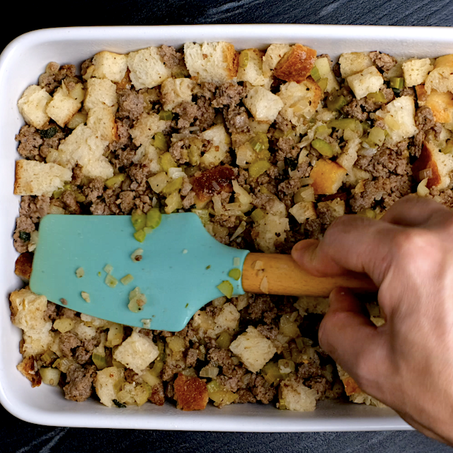 Cornbread sausage stuffing being pressed into a baking dish using a spatula.